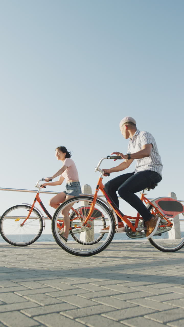 Vertical video of happy biracial couple riding bikes on promenade, in slow motion