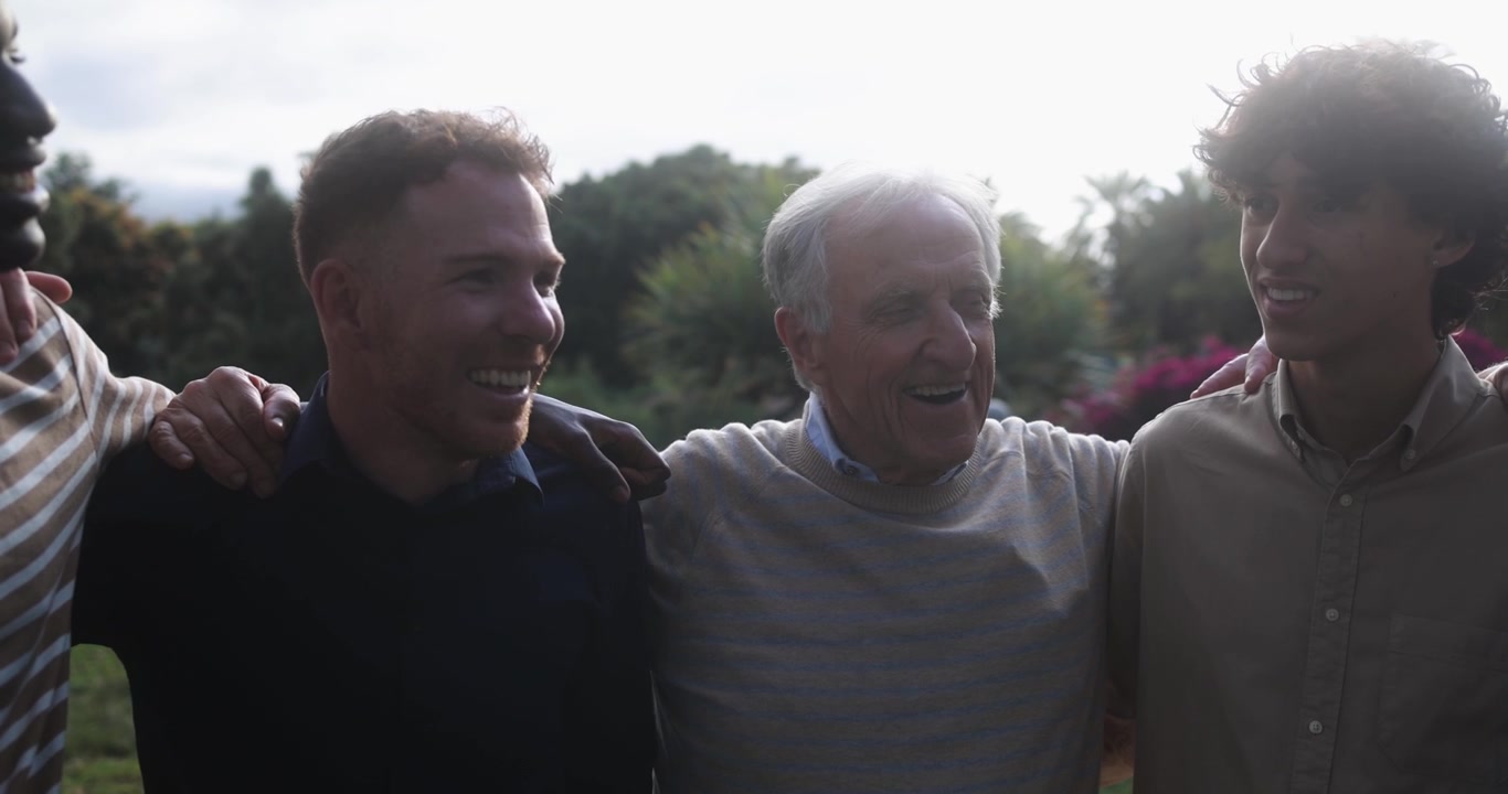 Glade mænd fra flere generationer, der griner og snakker sammen udendørs.