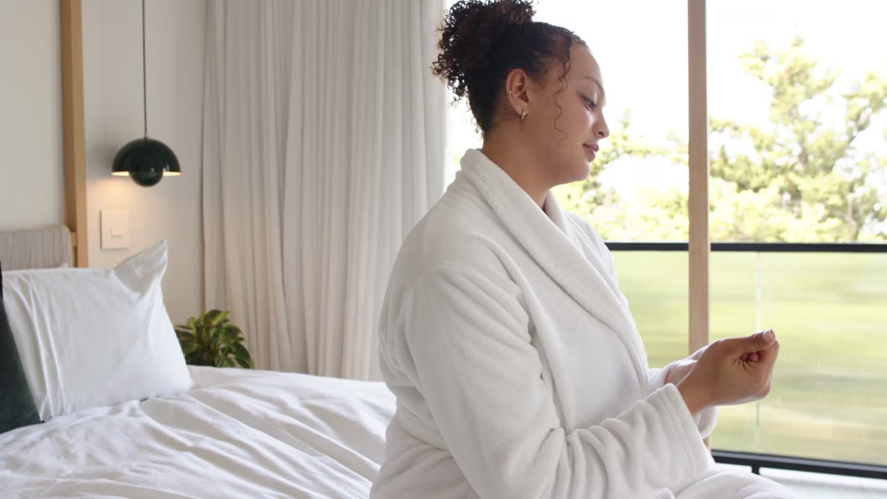 Donna afroamericana seduta sul letto in accappatoio, la luce del sole attira lo sguardo verso il balcone, mentre si accarezza e riflette.