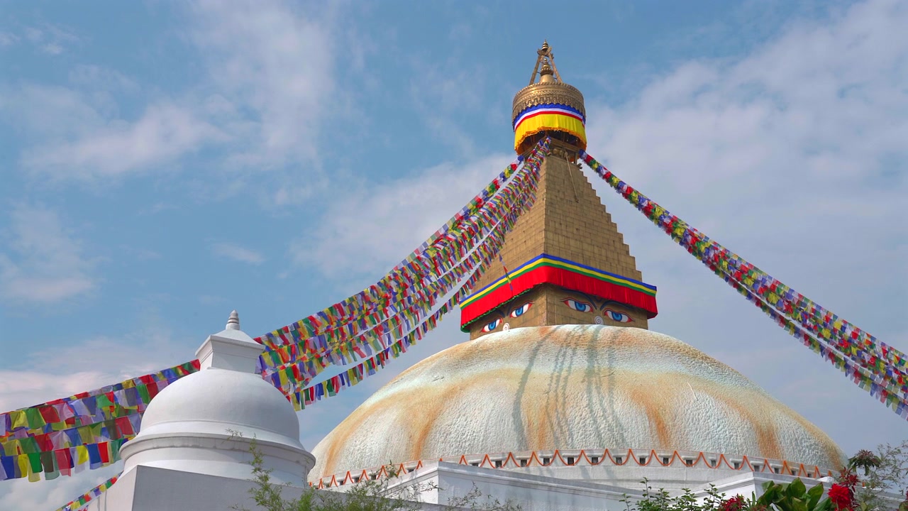 Staattinen keskikuvakulma suuresta Boudhanath Stupasta, jossa etualalla on pienempi valkoinen kupoli, koristeltuna värikkäillä rukouslipuilla kirkkaan taivaan alla Nepalissa.