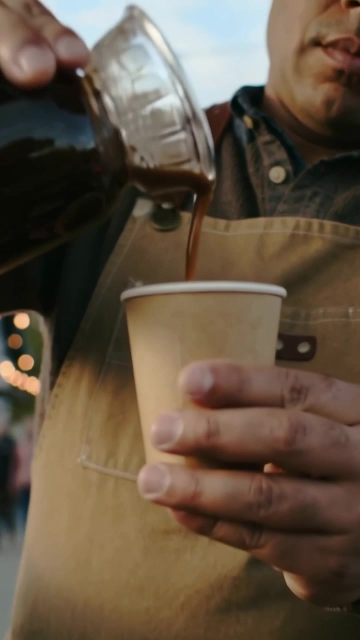 Vertikal video: En försäljare i förkläde ställer ner en pappersmugg och häller kaffe från en karaff till en gäst på en marknad.