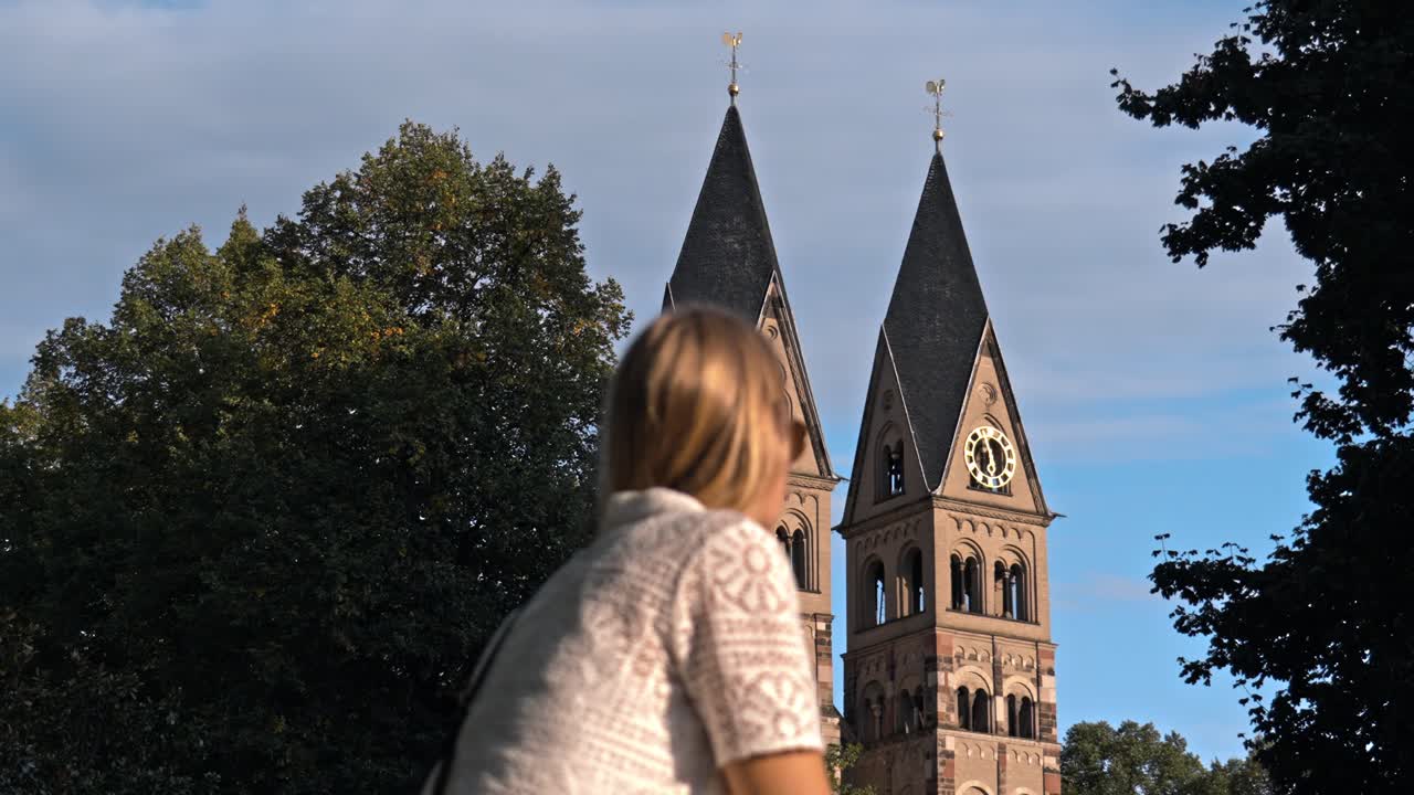 Blonde woman admiring the historic Basilica of St. Castor in Koblenz, Germany, with its twin Romanesque towers and dark pyramidal roofs dominating the background