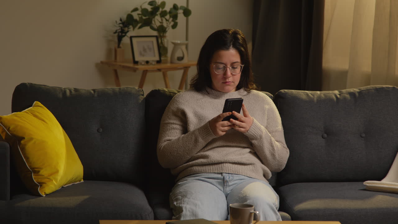 Young Woman Spending Evening At Home Sitting On Sofa With Mobile Phone Scrolling Through Internet Or Social Media 1