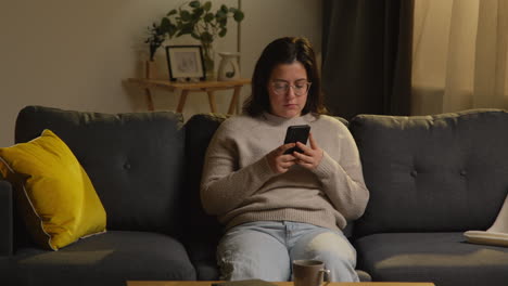 young woman spending evening at home sitting on sofa with mobile phone scrolling through internet or social media 1