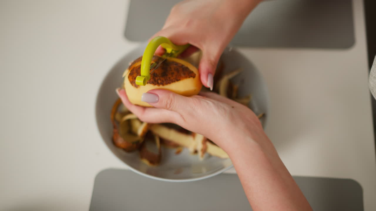 Close up aerial view of person peeling potato with green handle peeler above bowl filled with potato skin in home kitchen setting, focused on hands and tool with soft lighting over neutral background