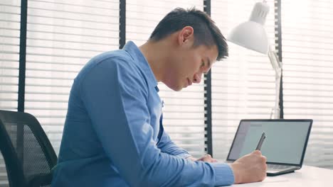 asian businessman use laptop computer analysing marketing in business. attractive handsome professional male employee worker sit on table, feel happy to write note on paper and working in workplace.