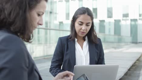 Colegas-De-Negocios-Femeninas-Enfocadas-Que-Usan-Una-Computadora-Portátil-Y-Una-Tableta-Para-Trabajar-Juntas,-Sentarse-Afuera,-Discutir-Contenido-Y-Escribir