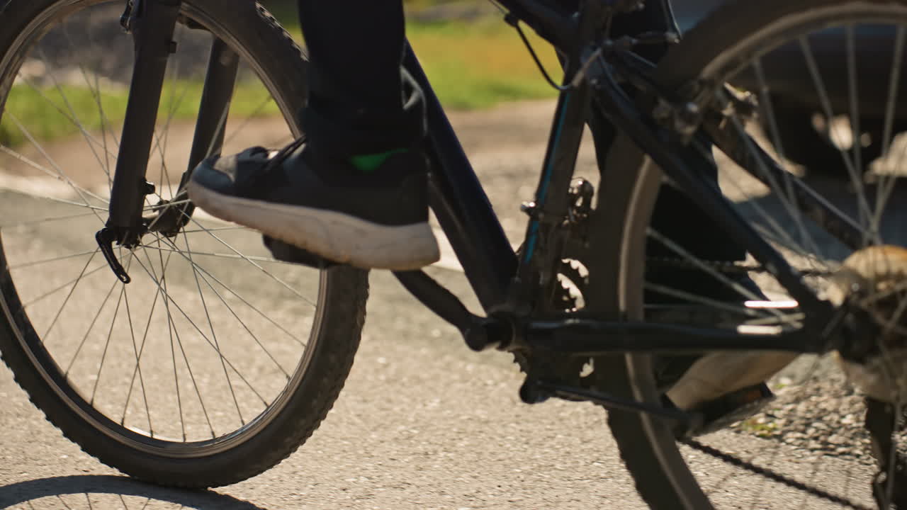 Close-up de pedal de bicicleta em área suburbana mostrando um ciclista branco. A luz quente do fim da tarde projeta uma longa sombra da roda sobre o asfalto liso, com detalhes do tênis no pedal, raios giratórios e ritmo constante.