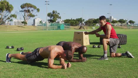 two diverse fit men exercising outdoors, doing plank while male trainer times them