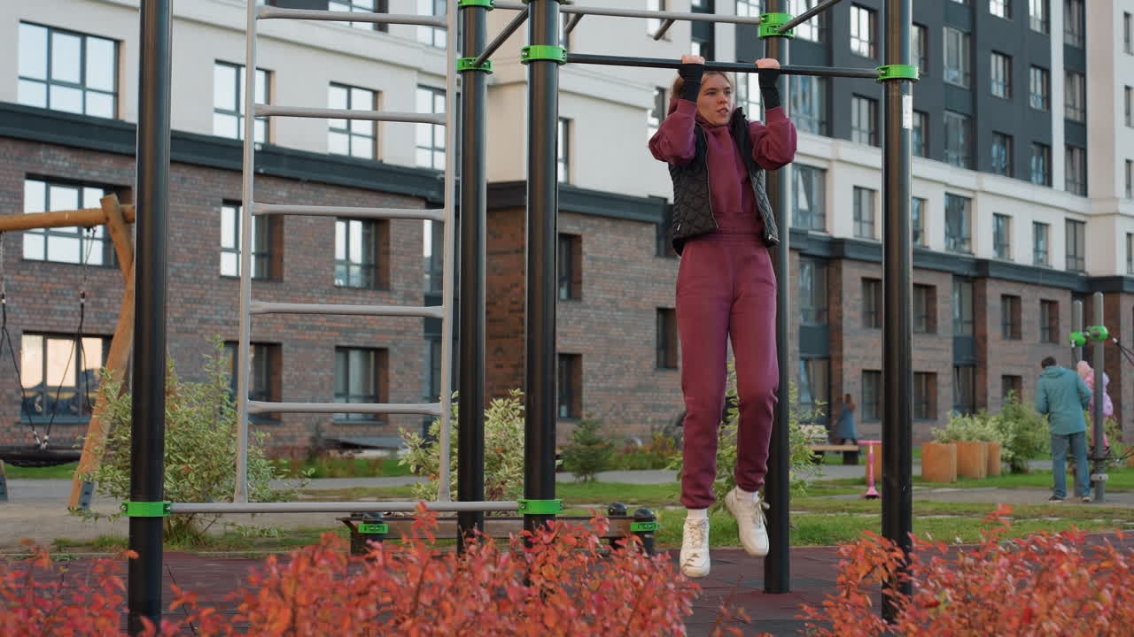 Energetic lady working out on horizontal pull up bar in urban park with people around on rubber flooring showcasing strength control focus during intense calisthenics exercise