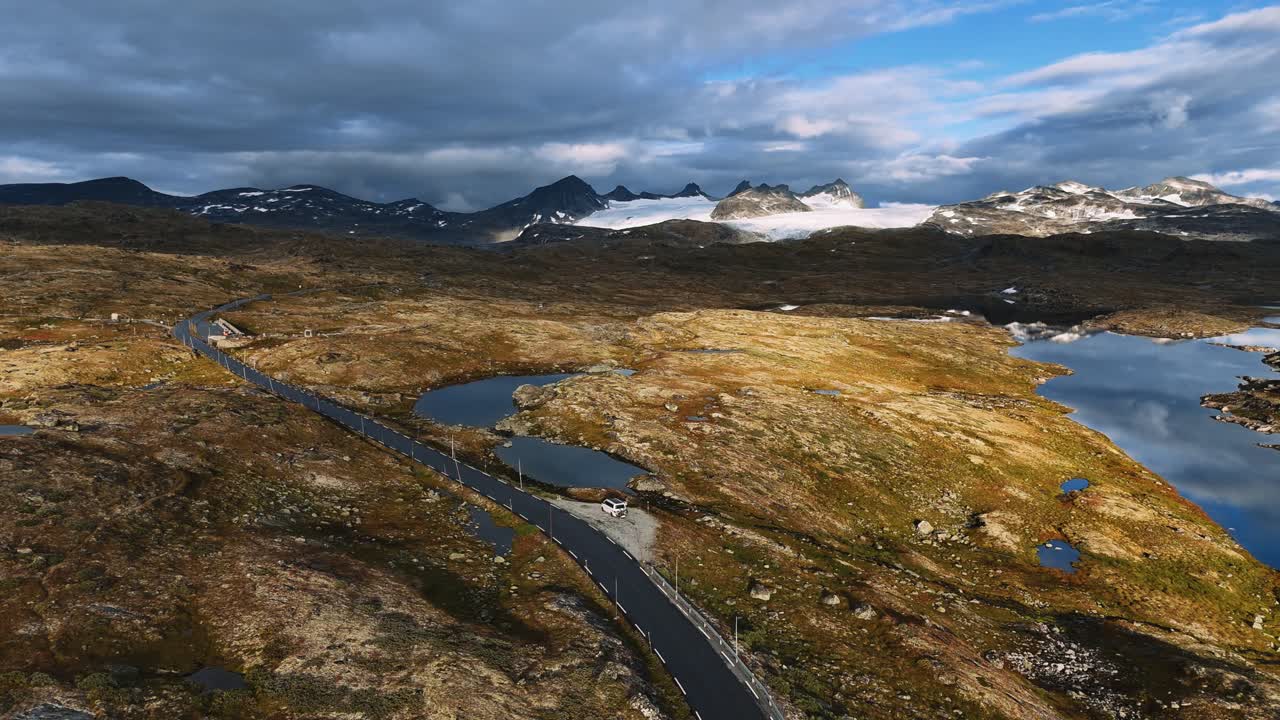 Valkoinen matkailuauto pysäköitynä autiolle ylänkö-/vuoristotielle jäätikköhuippujen ja tundrajärvien keskellä, Norja, ilmakuva.