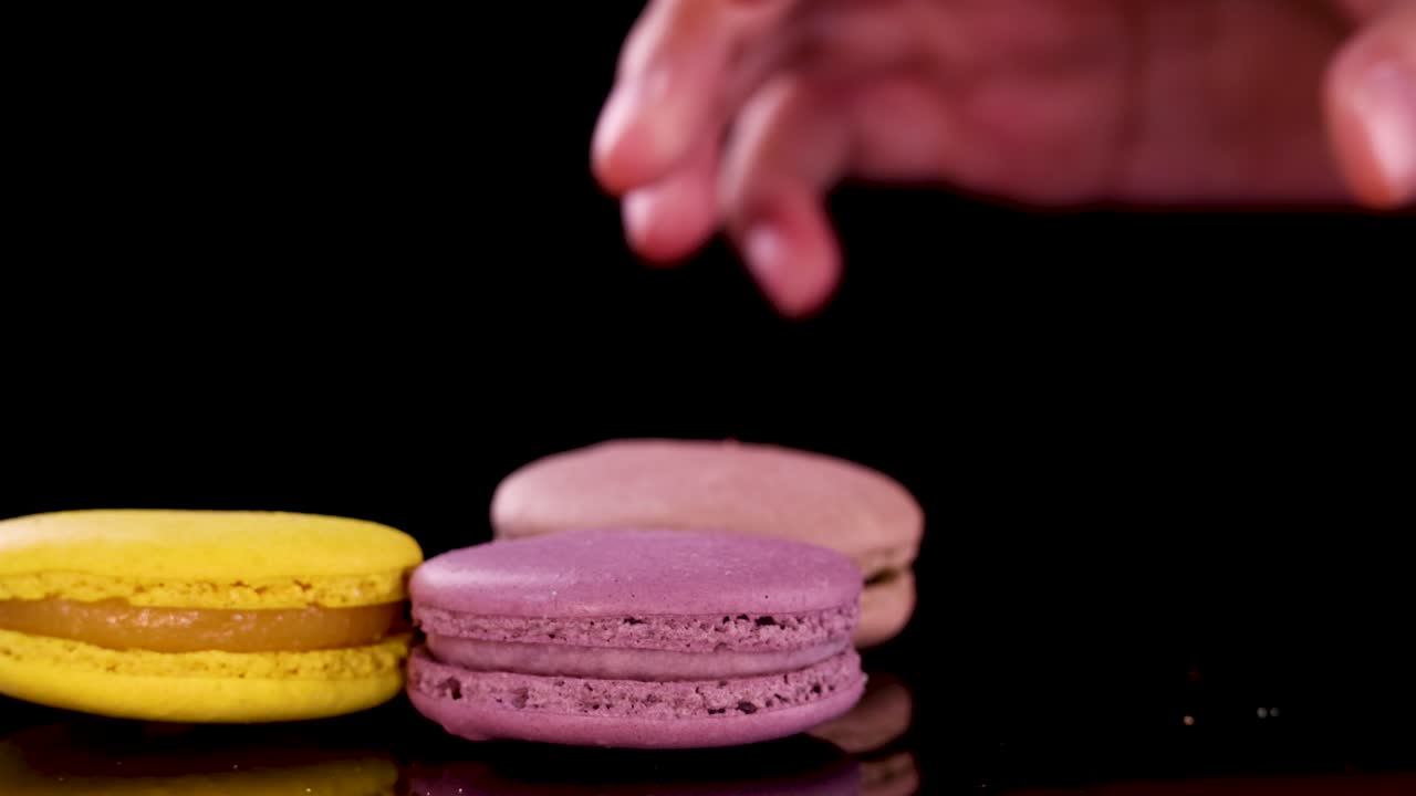A hand carefully picks assorted colorful macarons from a glossy black surface under bright, even lighting, highlighting the vibrant desserts in a close-up, static shot