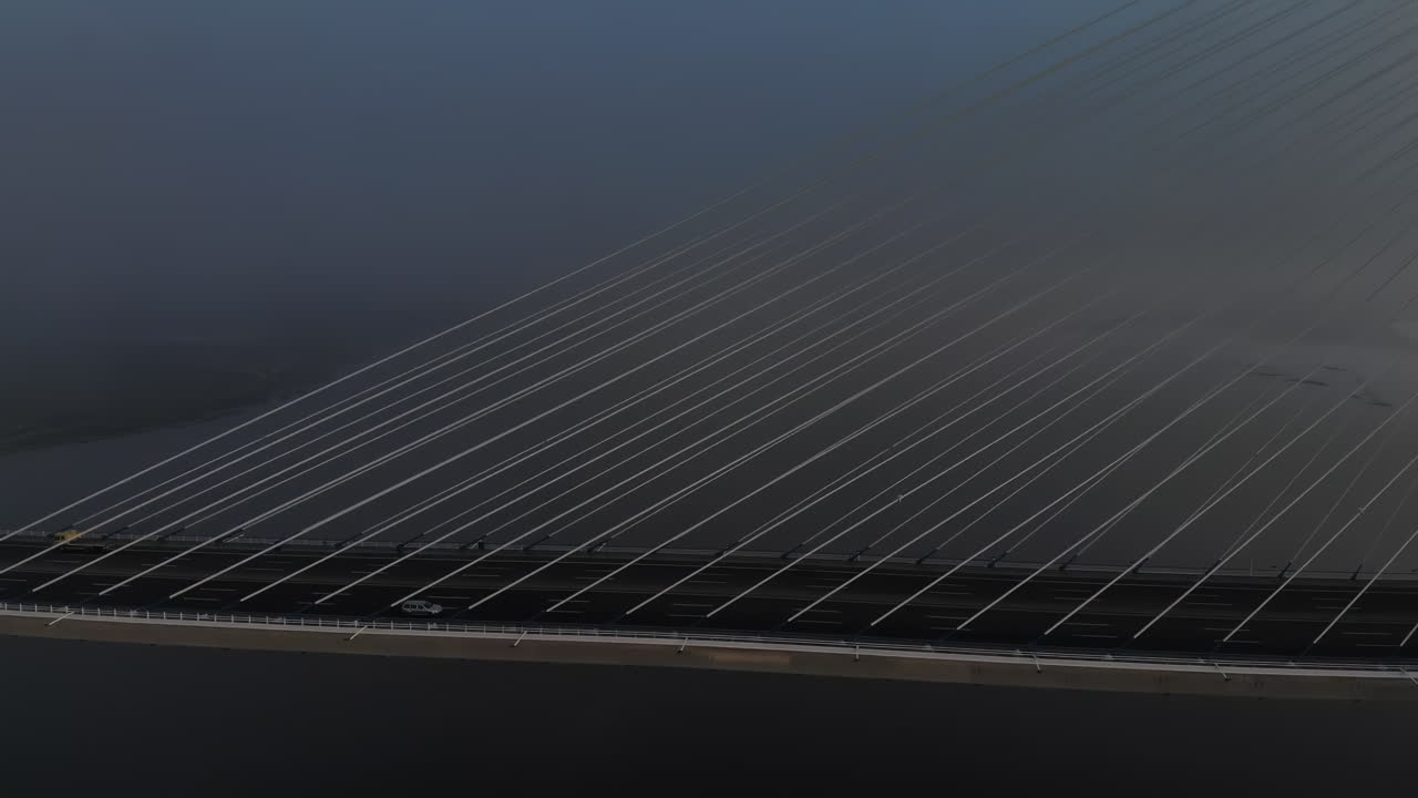 A breathtaking view captures a cable-stayed bridge shrouded in fog, with vehicles quietly moving across the structure during the early morning hours. The atmosphere is calm and mysterious