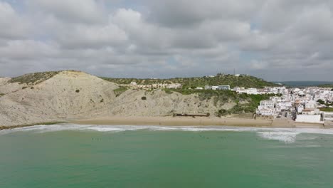 Toma-Aérea-Acercándose-A-Los-Surfistas-Esperando-Olas-A-Lo-Largo-De-La-Playa-De-Un-Pintoresco-Pueblo-Costero-Con-Casas-Blancas