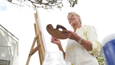 Feliz-Mujer-Mayor-Caucásica-Pintando-Cuadros-Y-Sonriendo-En-Un-Jardín-Soleado,-Cámara-Lenta,-Espacio-De-Copia