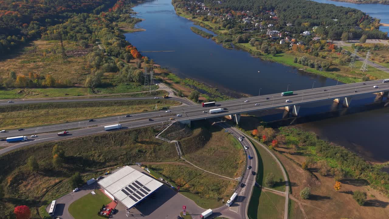 Aerial view of a highway bridge over the Nemunas River in Kaunas, Lithuania, surrounded by vibrant autumn landscapes, roads, and a roundabout near a gas station