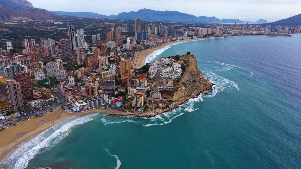 La costa di Benidorm dall'alto mostra un promontorio roccioso che divide due spiagge sabbiose, acque turchesi del Mediterraneo, uno skyline di grattacieli e uno sfondo montuoso sulla Costa Blanca spagnola.