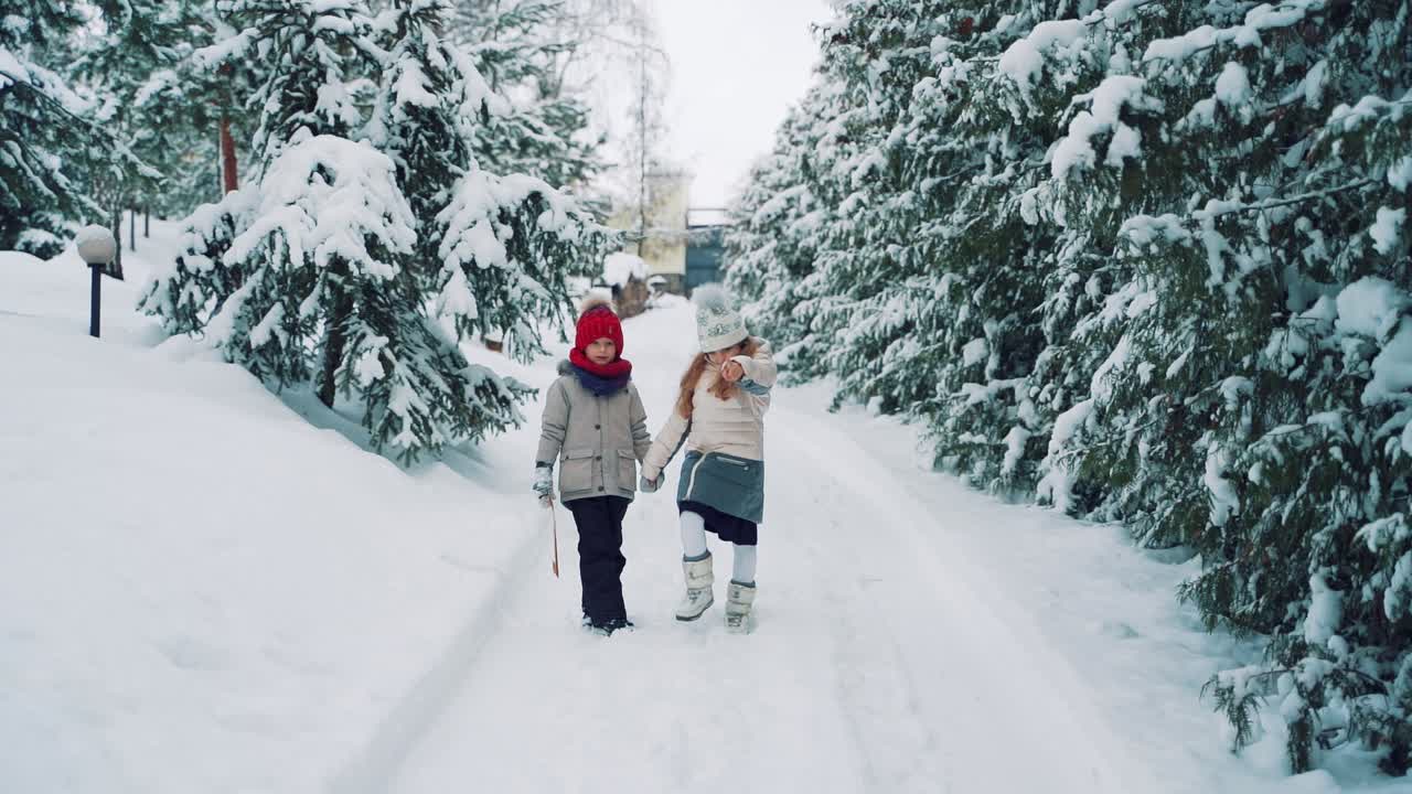 A little girl and a boy in warm clothes are walking along a snowy path holding each other's hands and talking on the background of green fir-trees. The boy is holding an envelope in his hands and listening to the story of a cute girl. Slow motion