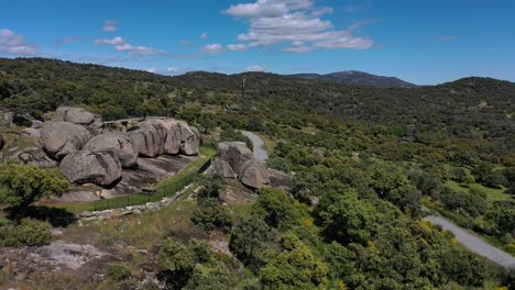 Orbital-flight-with-a-drone-over-a-viewpoint-located-on-a-large-granite-boat-and-rocks-around-it,-partly-fenced-by-a-stone-wall-and-metal-fence,-we-discovered-a-large-grove-of-Pelahustan-trees