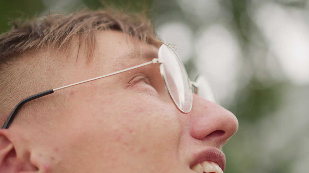 비가 오는 공원에서 렌즈에 물방울이 맺힌 채 안경을 쓰고 미소짓는 젊은 백인 남성의 클로즈업 프로필, 부드러운 보케 배경, 내성적인 분위기, 희망에 찬 학생의 느낌, 영화 같은 얕은 심도와 자연스러움