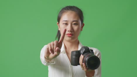 심각한 아시아 사진작가, 그녀의 손에 카메라를 들고 스튜디오에서 녹색 화면 배경에 서있는 동안 지표 손가락 표시 없이 불쾌감을 표합니다.
