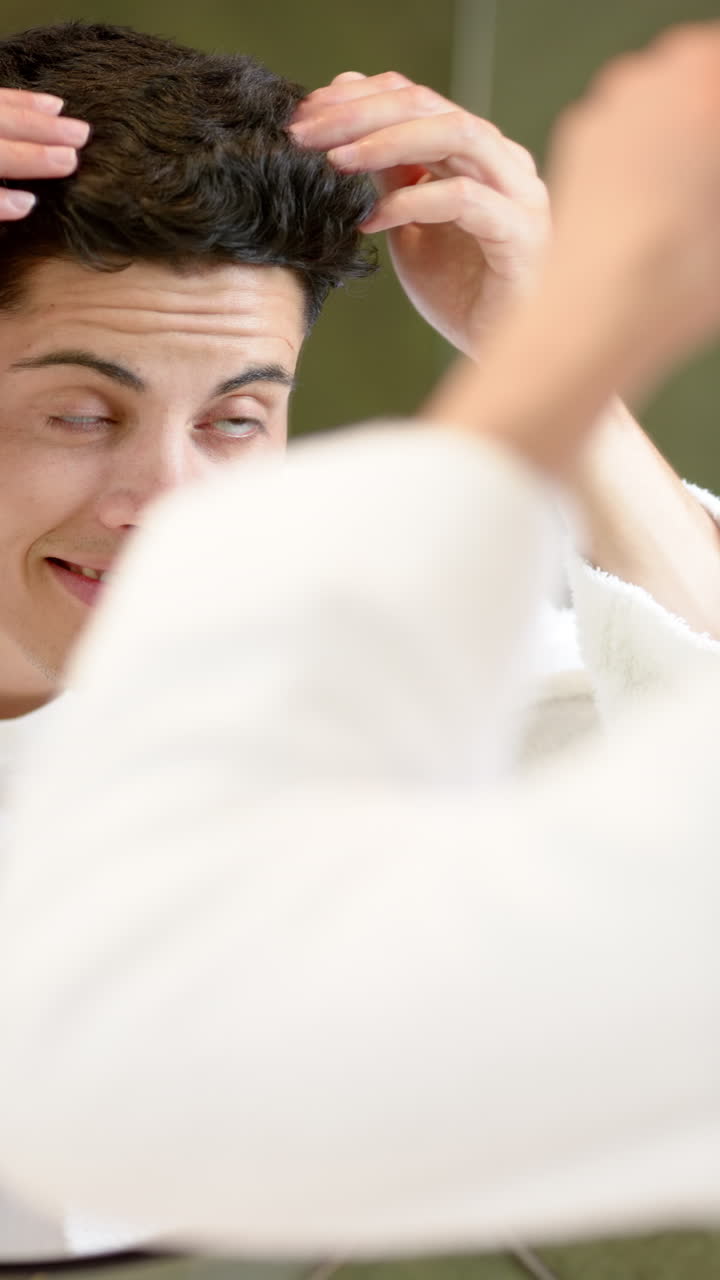 Vertical video of happy biracial man inspecting hair in bathroom mirror, slow motion