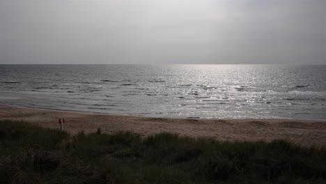 soft waves roll onto a swedish beach under an overcast sky. in slow motion, diffused light from a hidden sun shimmers on the water, creating a dreamlike, muted glow over the tranquil shoreline.