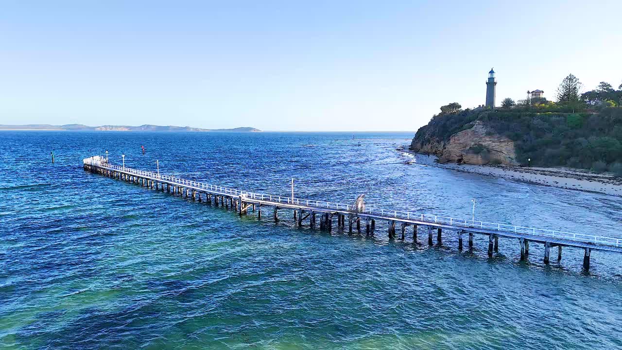 Aerial footage captures the serene Queenscliff Pier and lighthouse against a vibrant blue sea under clear skies