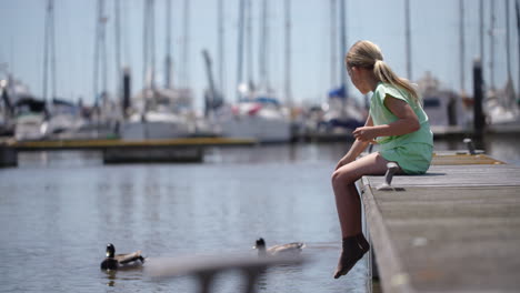 young girl sitting on wooden pier, dangling feet while feeding ducks near calm water during bright summer day, enjoying peaceful moment at scenic waterfront location