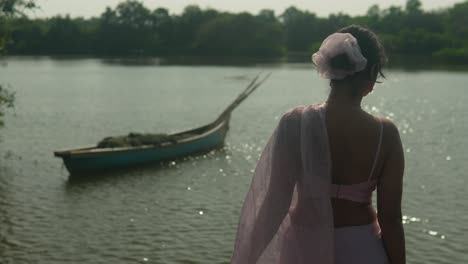 Woman-in-white-dress-by-tranquil-river-with-boat,-reflecting-sunlight-on-water,-serene-setting