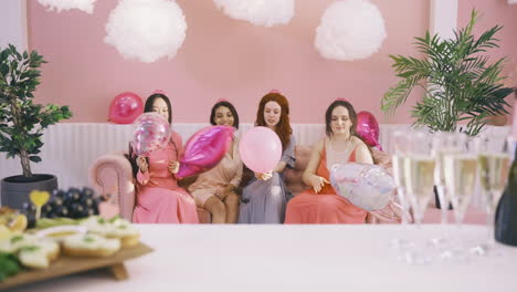 vista frontal de cuatro mujeres con tocados sosteniendo globos y hablando sentadas en el sofá
