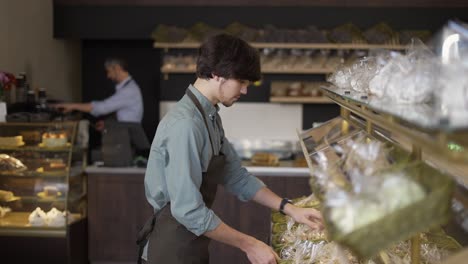 padre e hijo trabajando en una panadería familiar. el hijo endereza las mercancías en el estante mientras el padre enciende la máquina de café