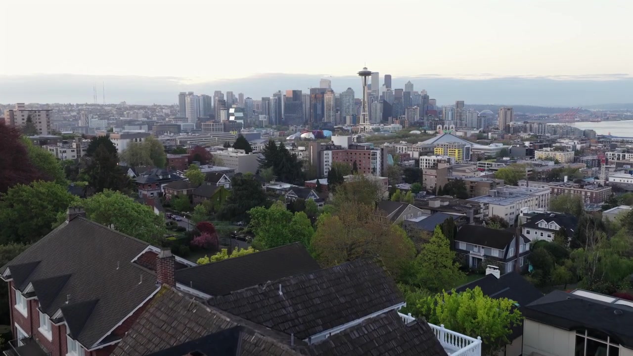 Residential rooftops and lush green trees fill foreground as Seattle’s iconic skyline with Space Needle stretches across Puget Sound at dusk over Seattle, Washington, USA. Aerial drone reveal wide