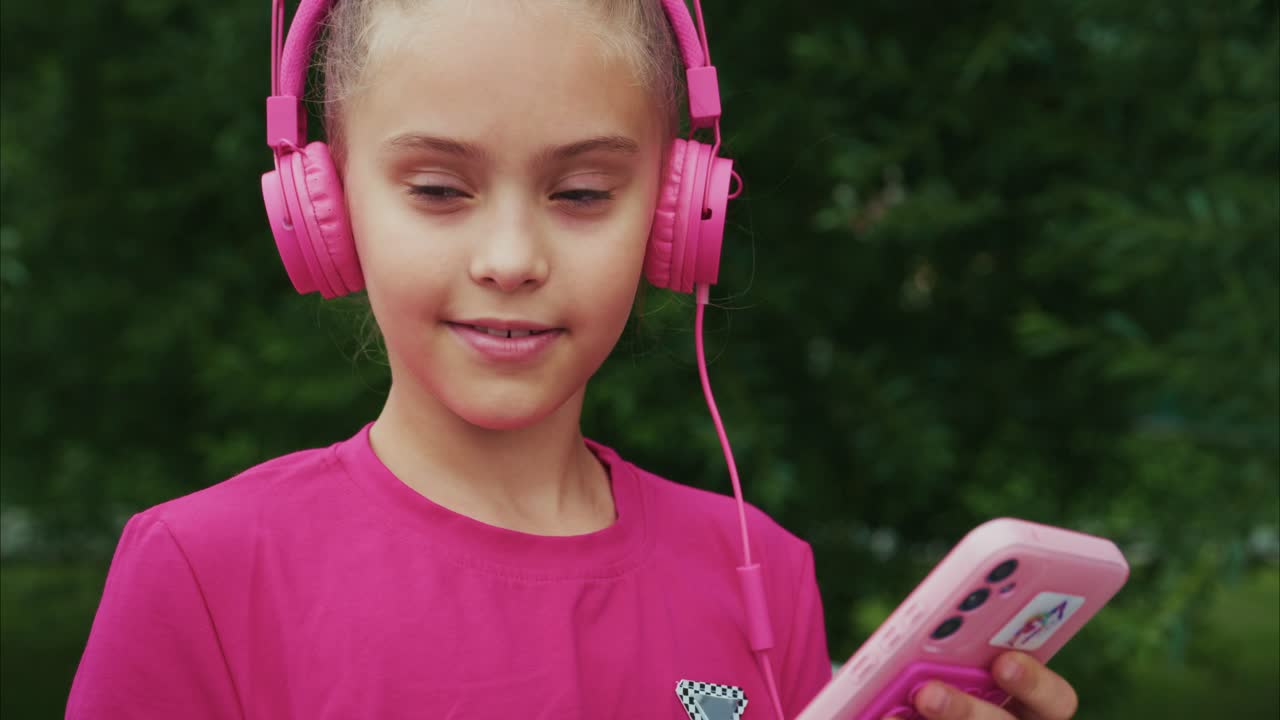 A young girl enjoying her music with vibrant pink headphones while attentively looking at her smartphone in a lush green outdoor setting