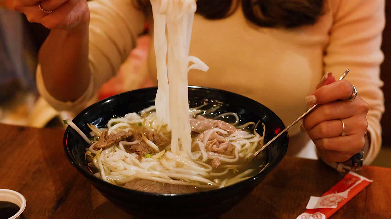 A person skillfully uses chopsticks to enjoy a steaming bowl of Vietnamese pho in a cozy restaurant setting