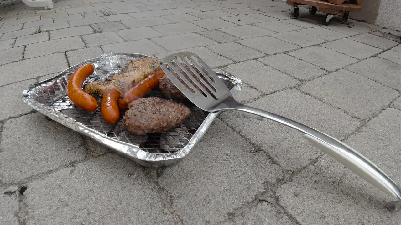 vista bloqueada de la mini parrilla de barbacoa desechable con diferentes carnes y espátula apoyada contra ella a cámara lenta