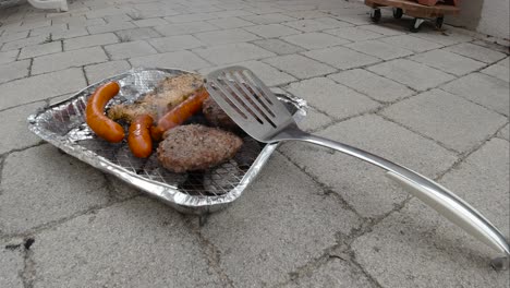 Vista-Bloqueada-De-La-Mini-Parrilla-De-Barbacoa-Desechable-Con-Diferentes-Carnes-Y-Espátula-Apoyada-Contra-Ella-A-Cámara-Lenta