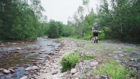 Wanderer-Mit-Rucksack-Spaziert-Mit-Seinem-Hund-Am-Felsigen-Fluss-Entlang