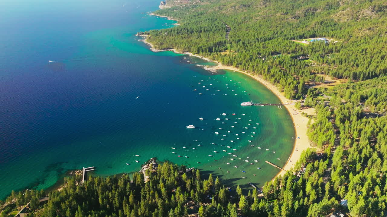 Flying closer to the numerous boats in the bay of Lake Tahoe. Pine trees overgrowing the shore. Aerial perspective on sunny day.