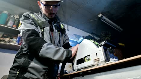 a young man with a beard in gray overalls by profession a carpenter works with a circular cutting machine