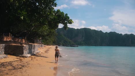 beautiful woman with her flip-flops in her right hand walks barefoot on the beautiful beach as small waves of the blue water wet her feet