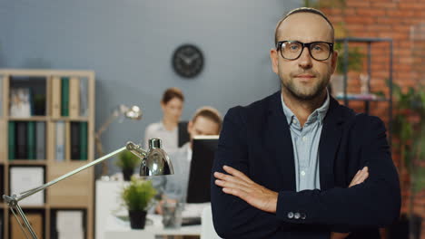 porträtaufnahme des gutaussehenden geschäftsmannes mit brille und offiziellem stil, der im büroraum steht, sich auf den tisch lehnt und den kopf zur kamera dreht