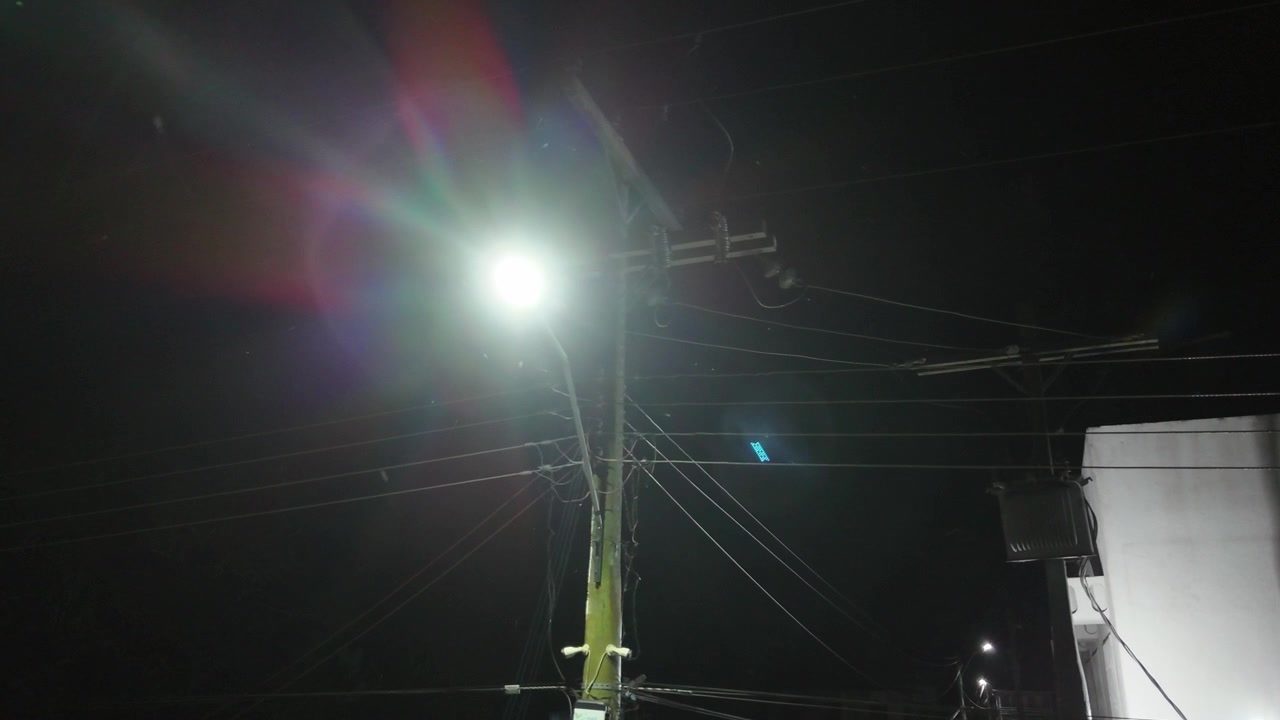 Illuminated street light against the night sky with utility pole and overhead power lines, captured in rural Brazil, handheld low angle view