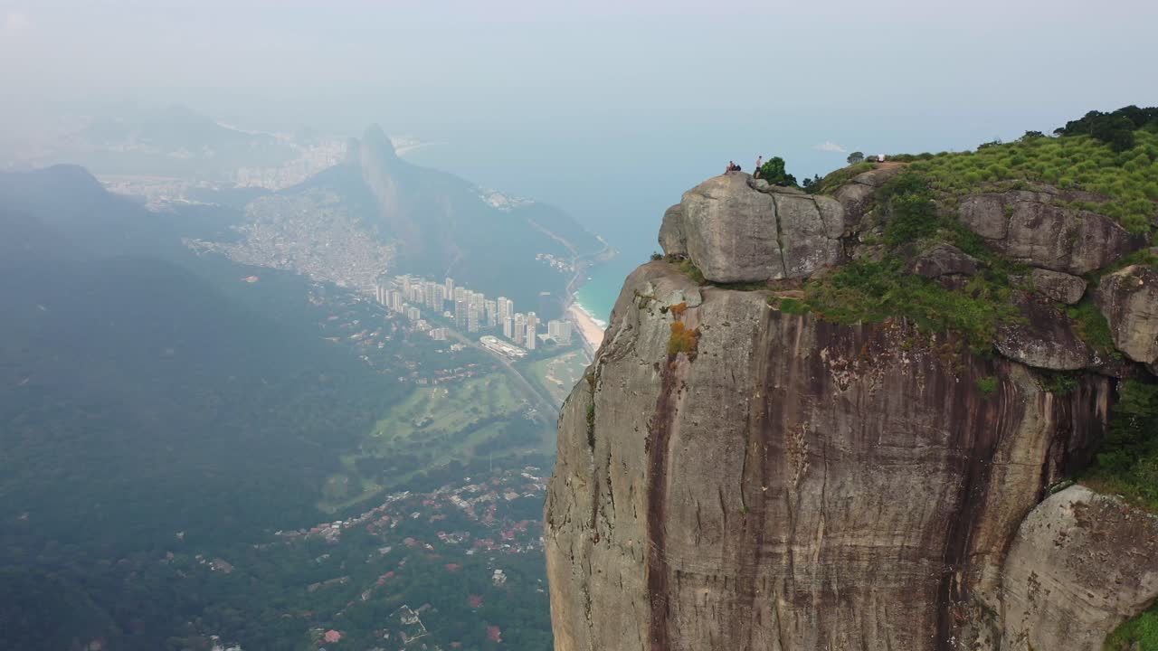 Kinoreifer Panoramablick über Rio de Janeiro von einem hohen Berggipfel, der Küstenlinie, Strände, Meer, Klippen und die Stadtsilhouette zeigt. Ein Reisender steht auf Felsen und blickt auf die ikonische brasilianische Landschaft