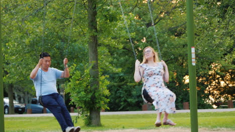 active young family resting in the park ride on a swing pregnant woman and asian man