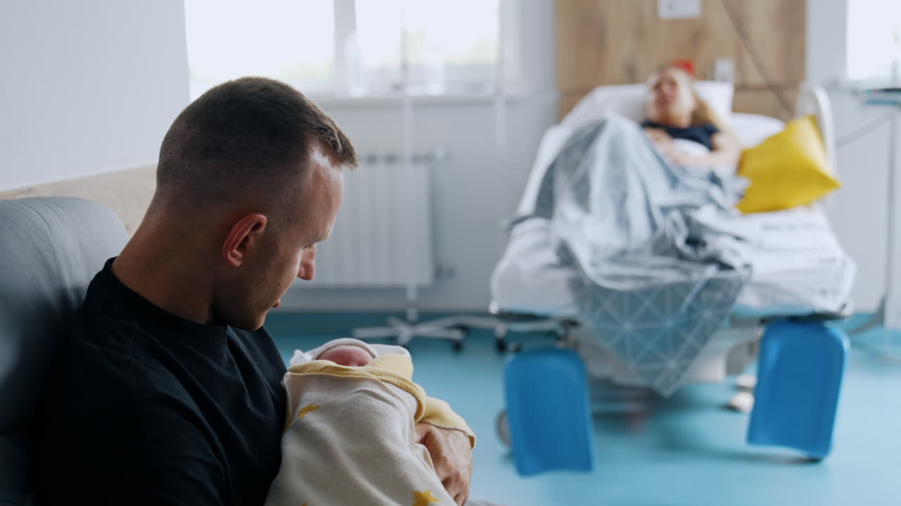 New-made father sitting and holding a newborn baby. Mother is resting in bed after childbirth at backdrop in blur.