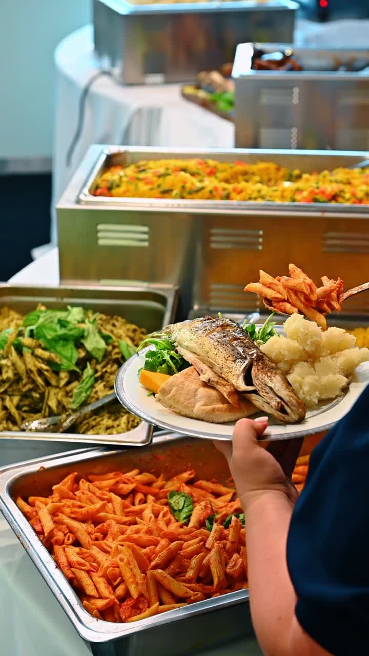 Persona che tiene un piatto con pesce grigliato e patate al buffet. Primo piano di una mano che tiene un piatto di ceramica bianca con pesce intero grigliato, purè di patate, pane pita e verdure.