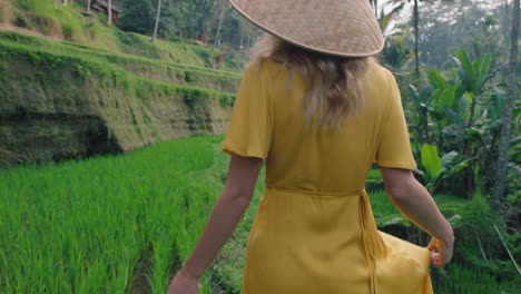 woman walking in rice paddy wearing yellow dress with conical hat exploring lush green rice terrace in cultural landscape exotic vacation through bali indonesia discover asia