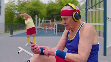 senior man after sport training listening music from mobile phone wearing headphones on playground