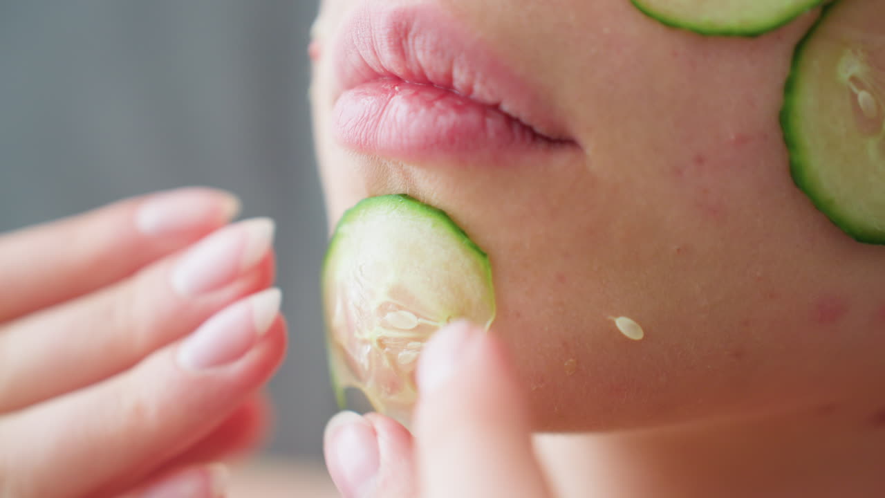 Close-up hand view of woman placing cucumber slices on her face for skin care, focused on the delicate placement of slices over her eyes and chin, showcasing self-care routine